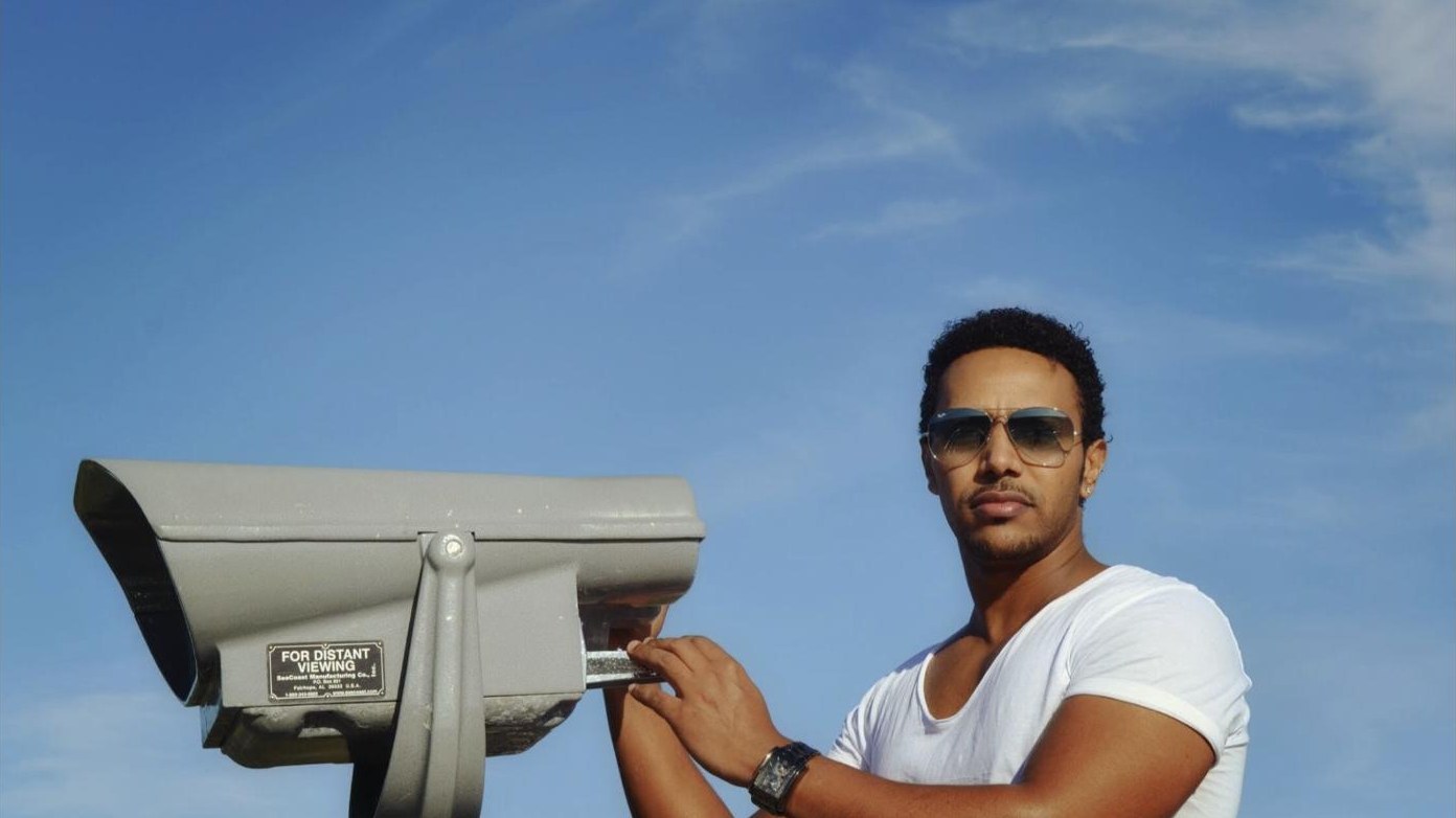 Milly Wessy in a white T-shirt and sunglasses beside a coin-operated viewfinder, under a blue sky